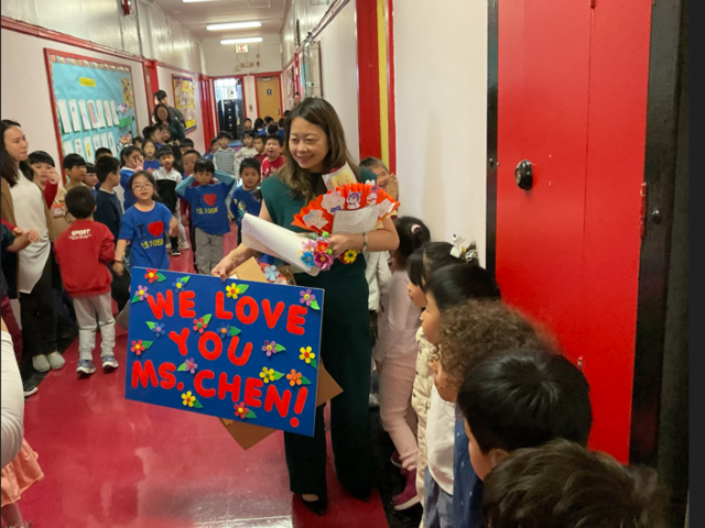 Crowd of children in a school hallway celebrating a teacher with flowers and a sign.