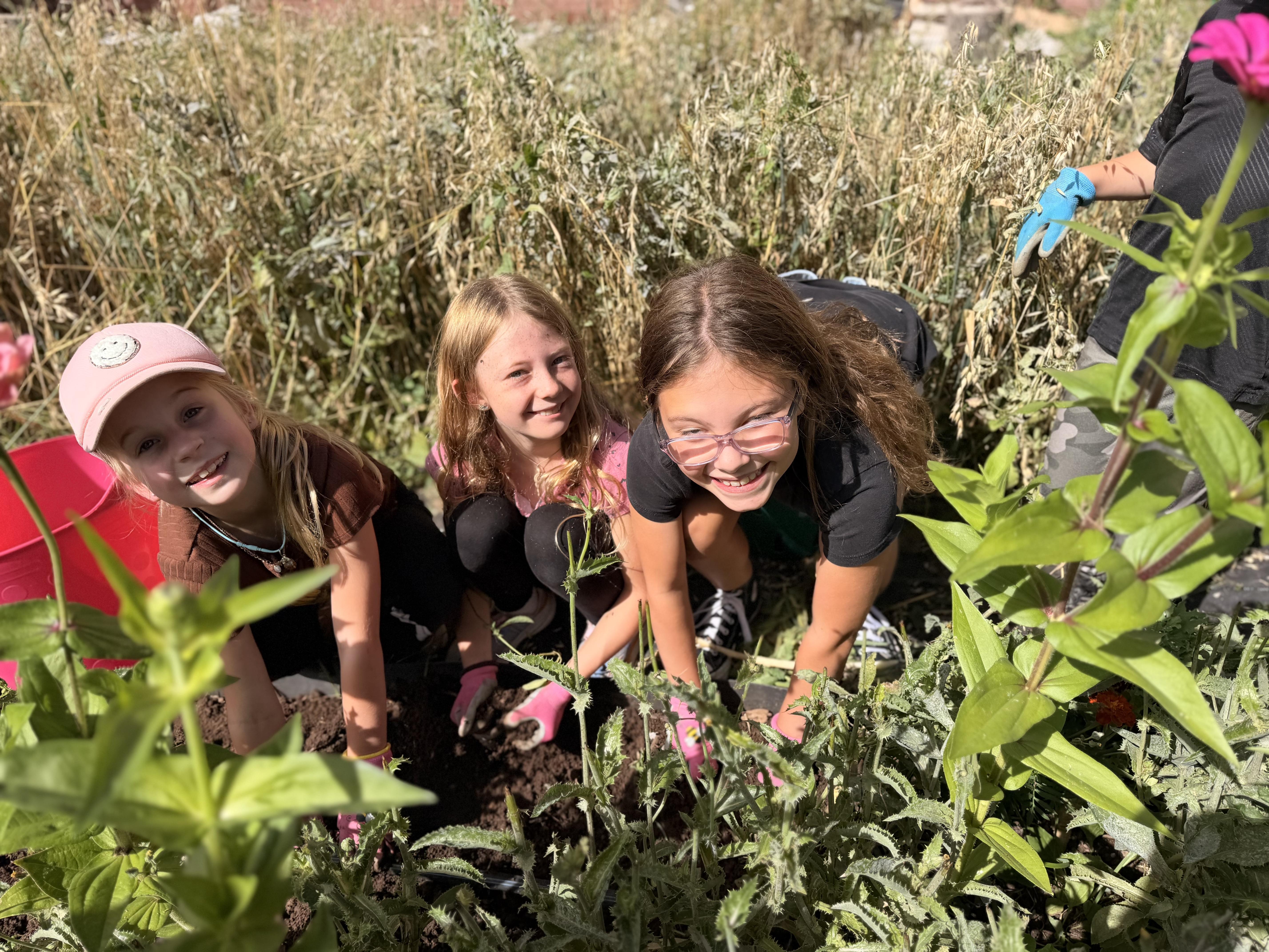 Three smiling children crouch together in a garden, helping tend to soil and plants.