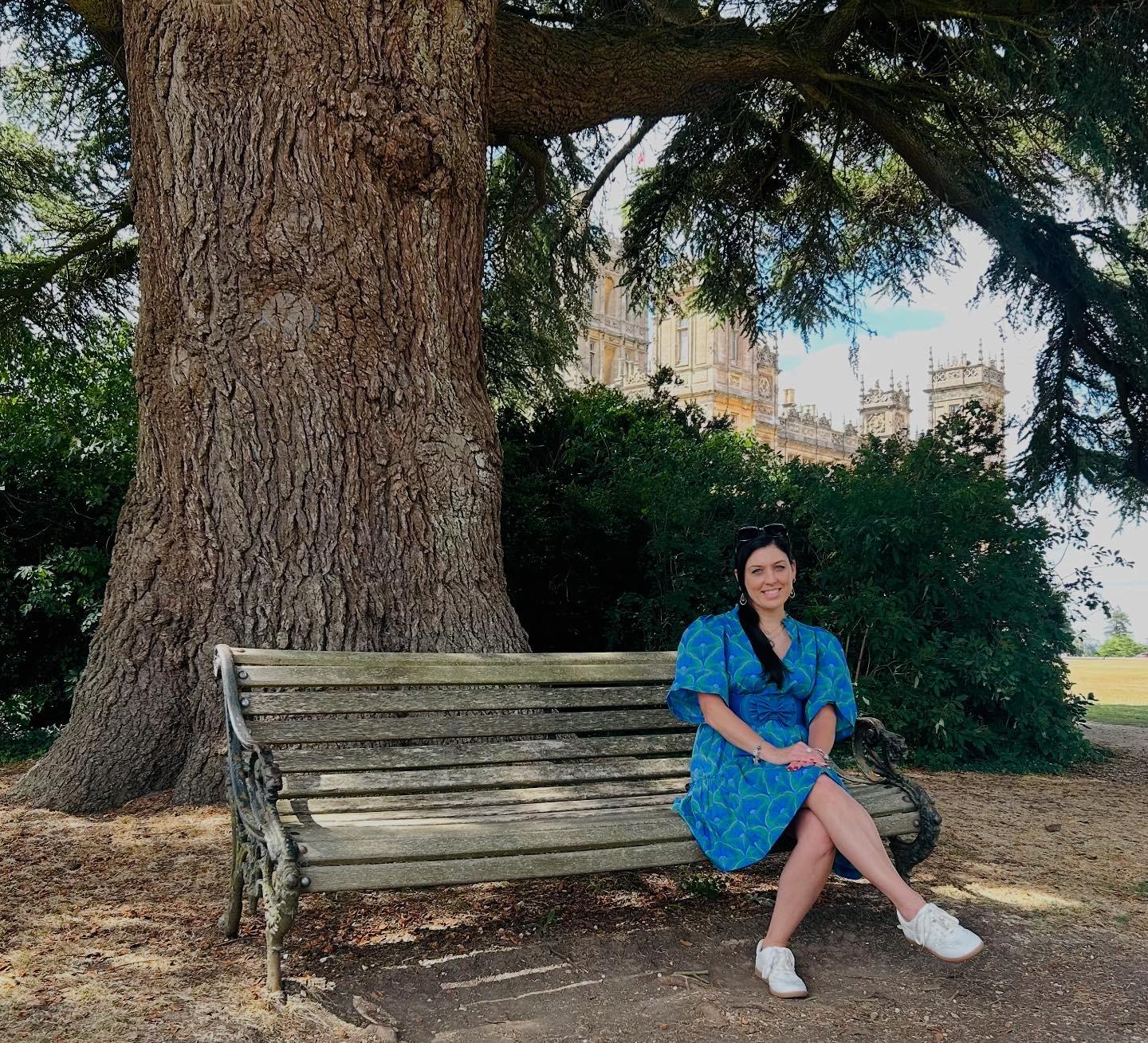 A woman in a blue patterned dress sits on a wooden bench by a large tree.