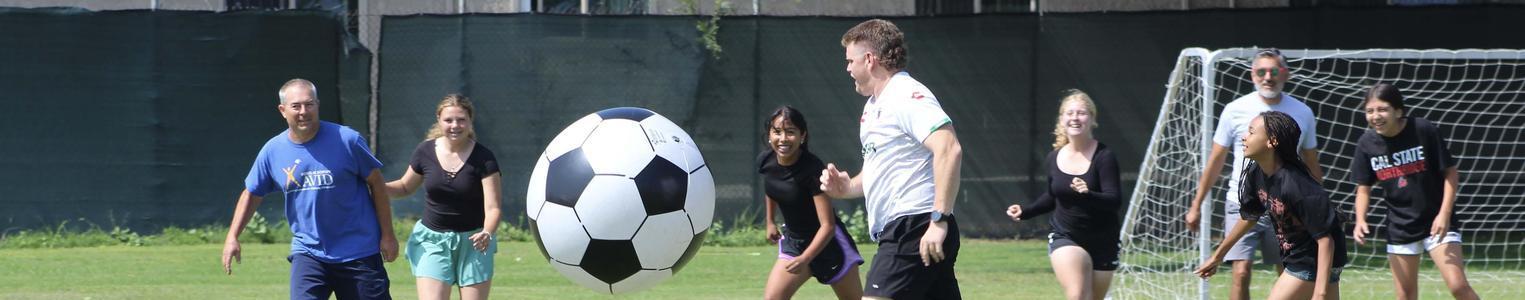 Adults and children participate in a soccer game with a large soccer ball.
