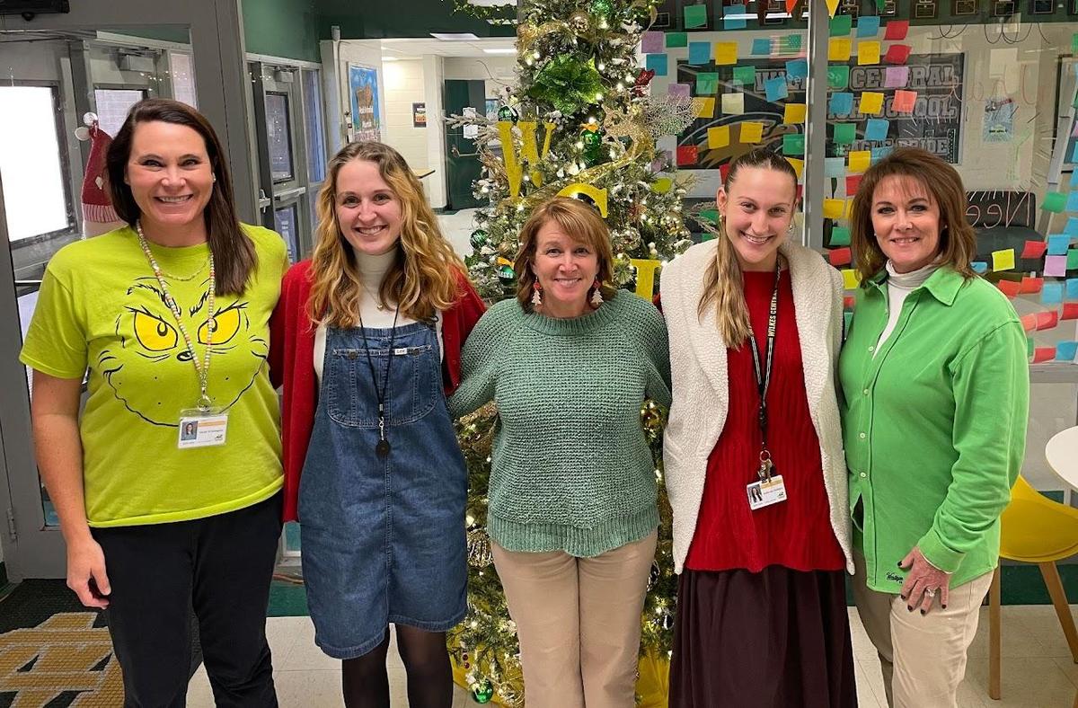 Five women smiling in front of a holiday tree decorated with colorful ornaments.