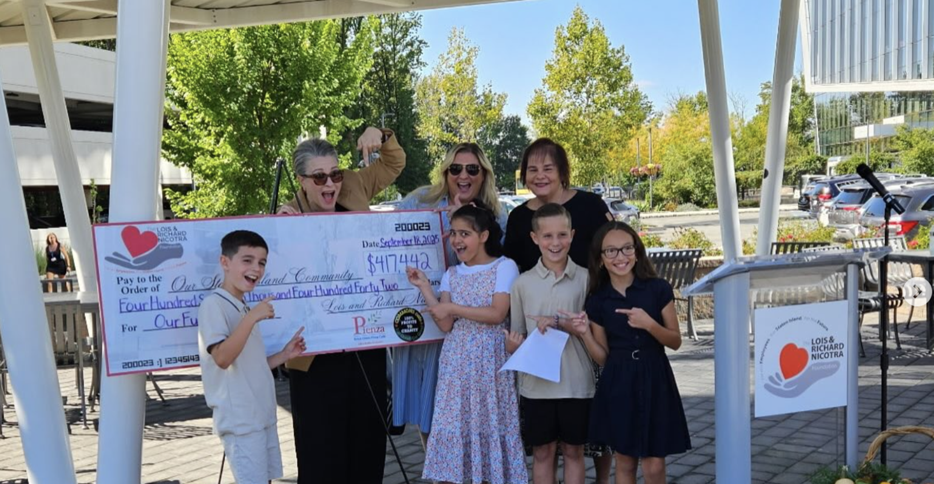 Group of people holding a large check outdoors, smiling and celebrating.