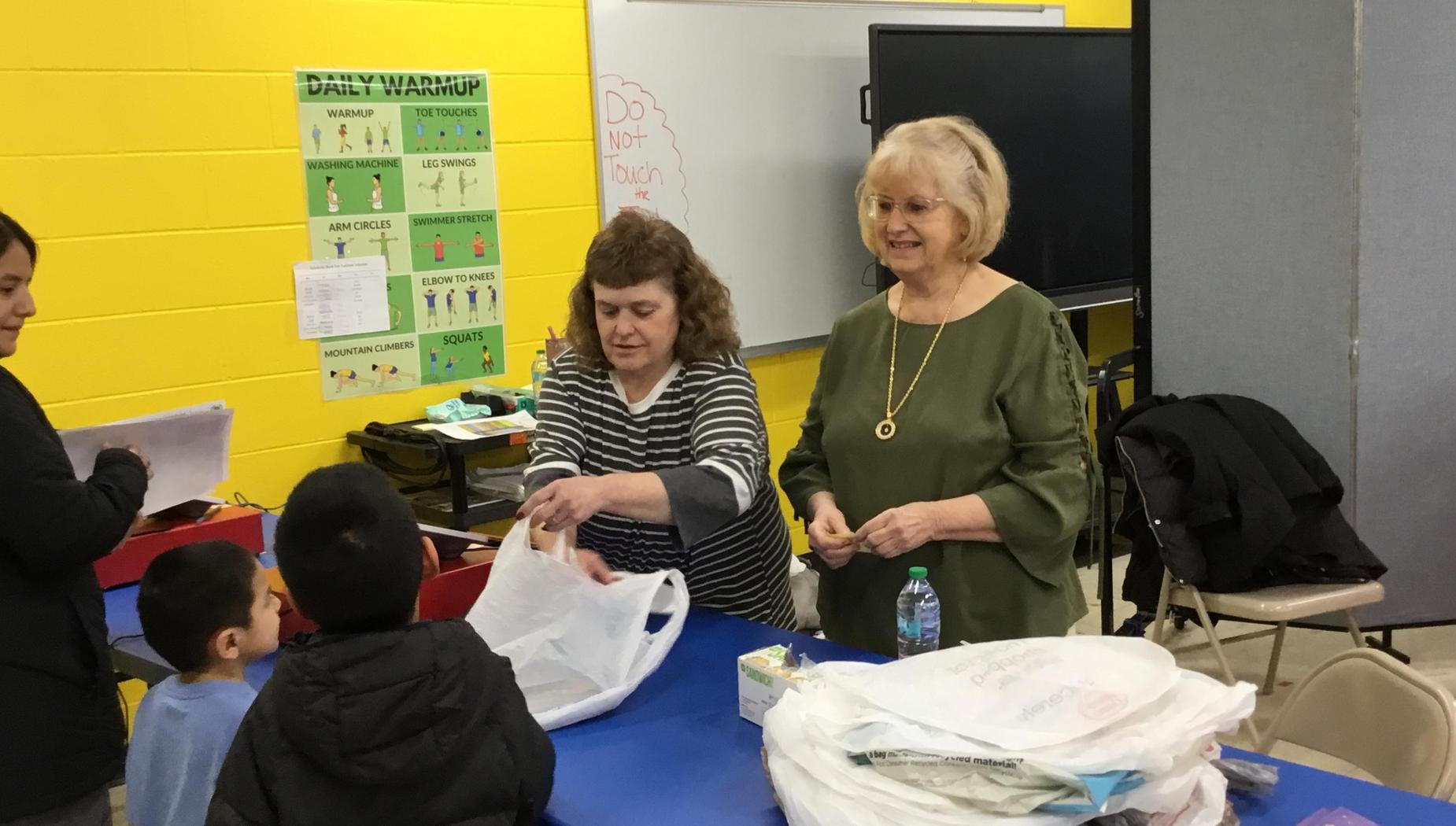 Two women organizing materials, with children present in a colorful classroom environment.