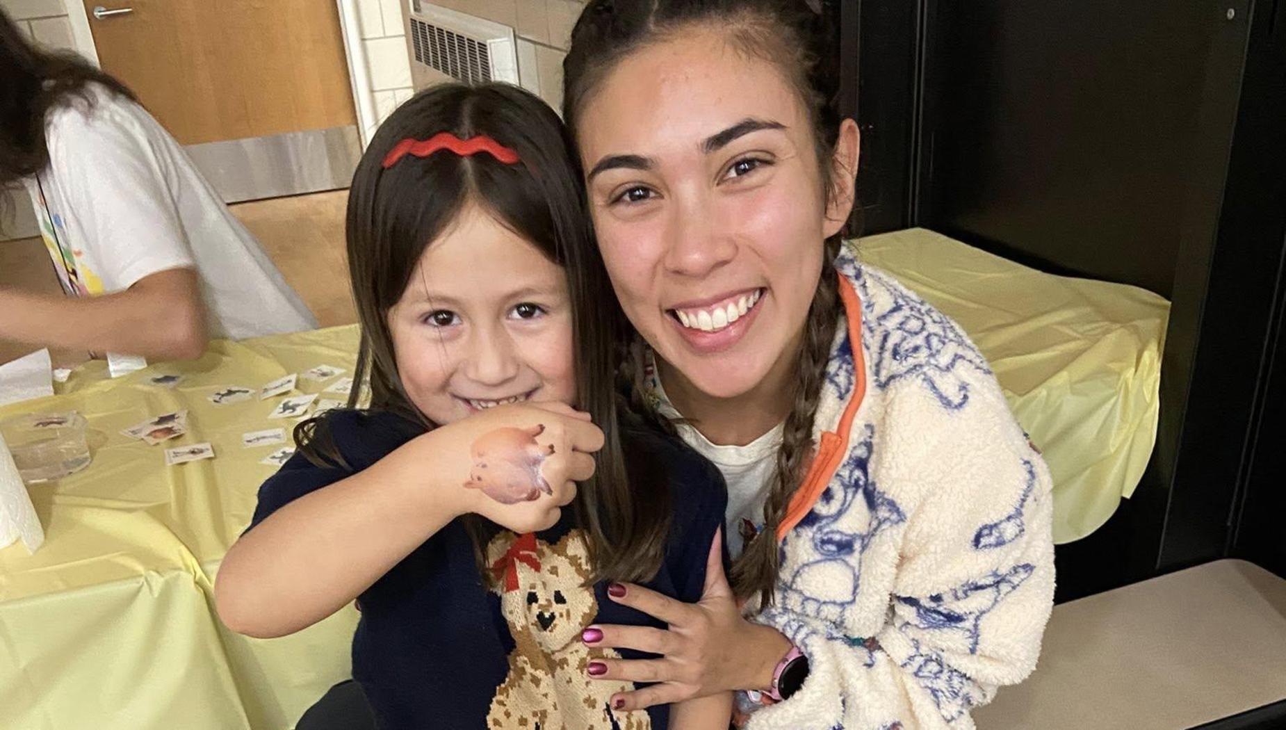 Two girls smiling together, one wearing a teddy bear sweater, in a cozy setting.