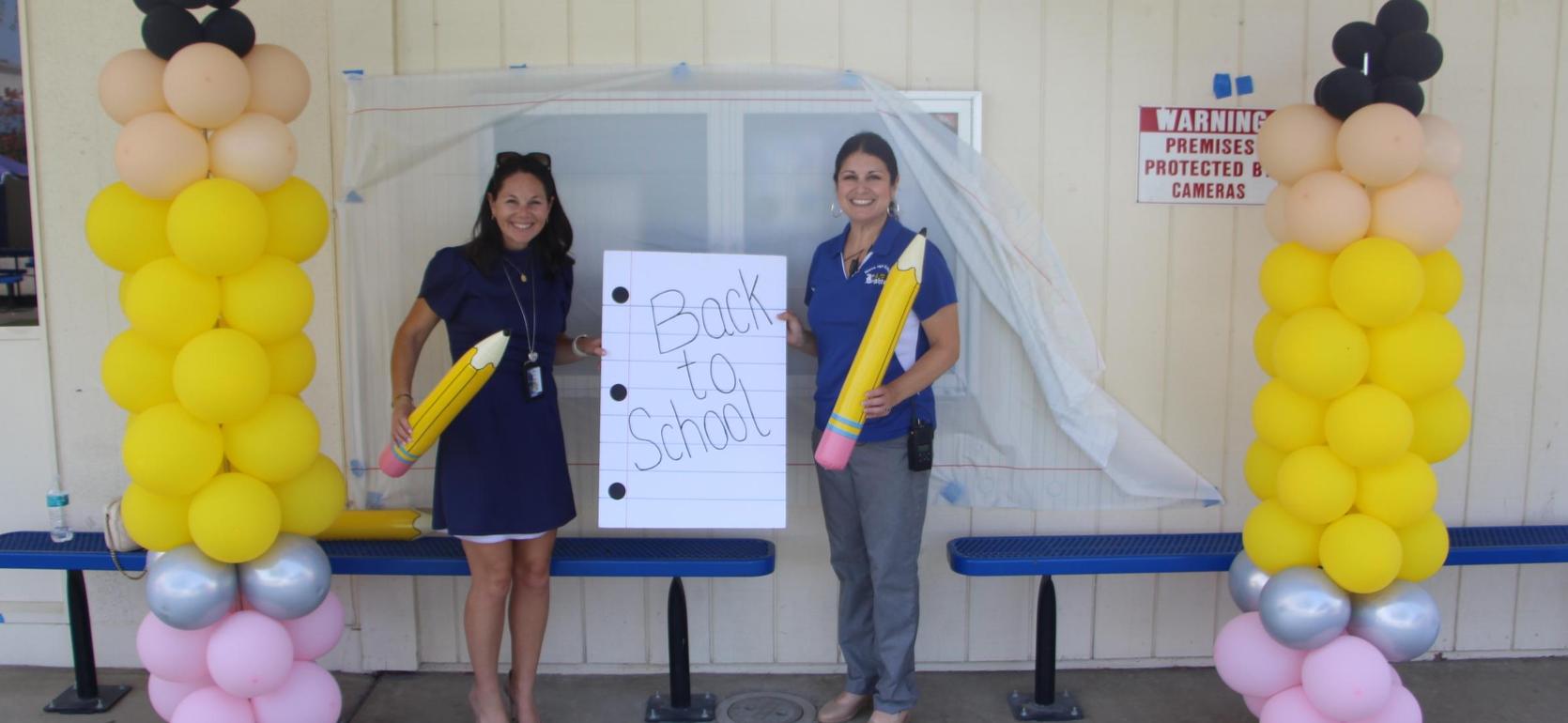 Two women holding a sign that says 'Back to School' with colorful balloon decorations.