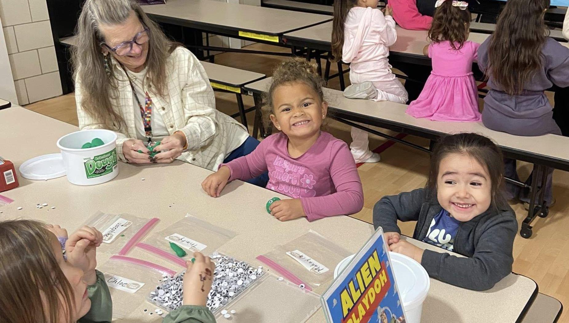 Children engaged in crafts at tables, with a woman assisting them and smiling.