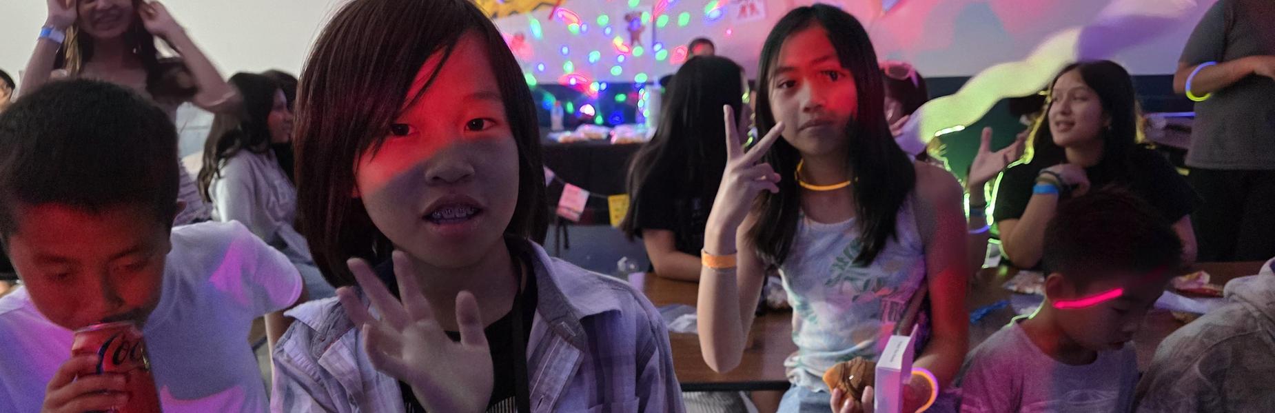 A group of kids with glow accessories enjoys a lively gathering, some waving at the camera.