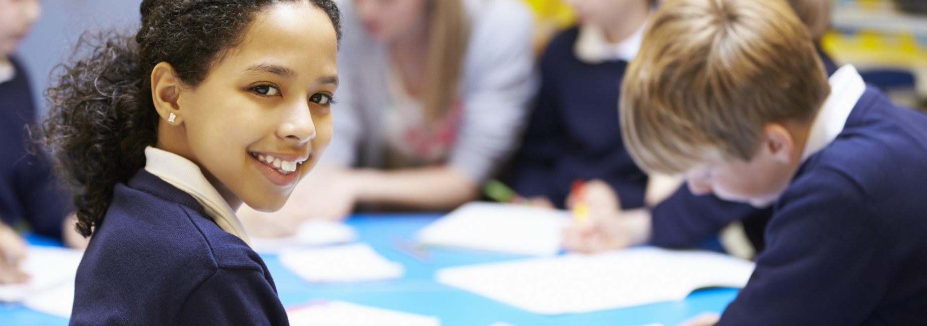 A girl in a navy sweater smiles at the camera while classmates are engaged in activities.