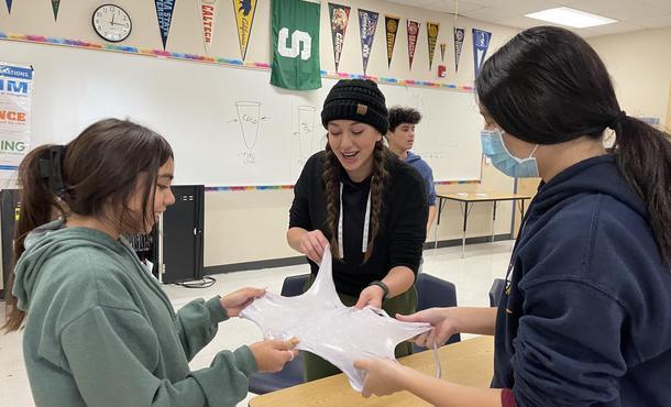 3 students smile and work together to stretch out slime to its thinnest property in a classroom with a collection of college pennants hung from the ceiling.
