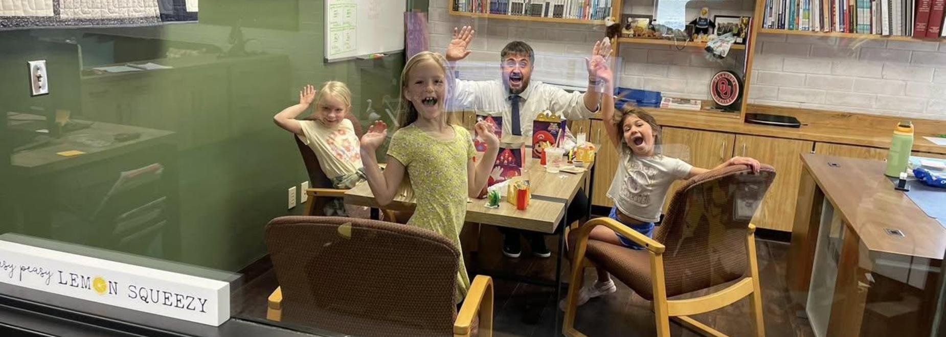 Four children and an adult smiling and waving from behind a glass wall.