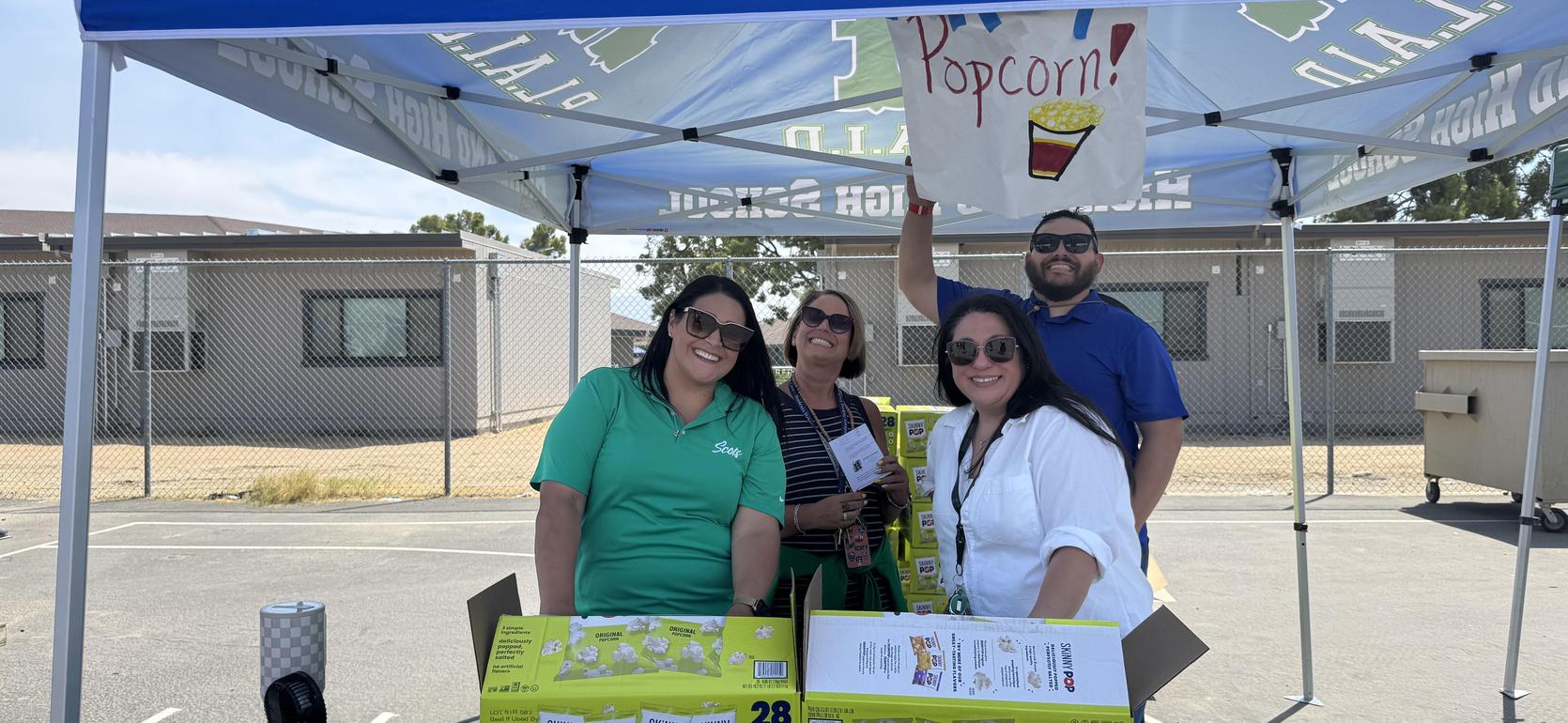 Four people stand under a tent, one holding a sign that says 'Popcorn.'