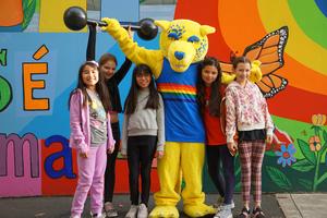 Acro Iris students pose with the school’s mascot, J.J. the Jaguar, in front of a colorful campus mural.