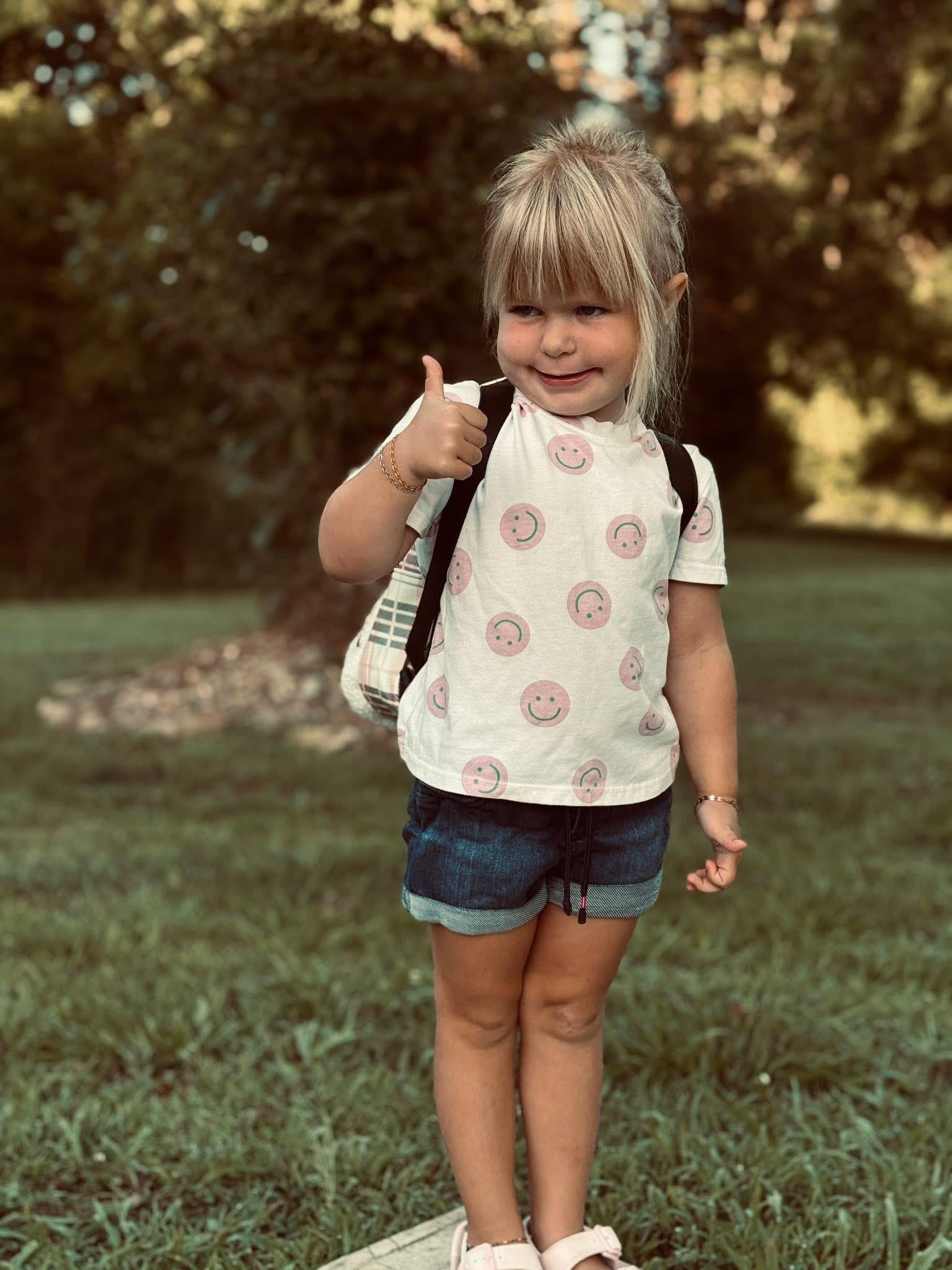 A smiling girl with a backpack gives a thumbs up outdoors.