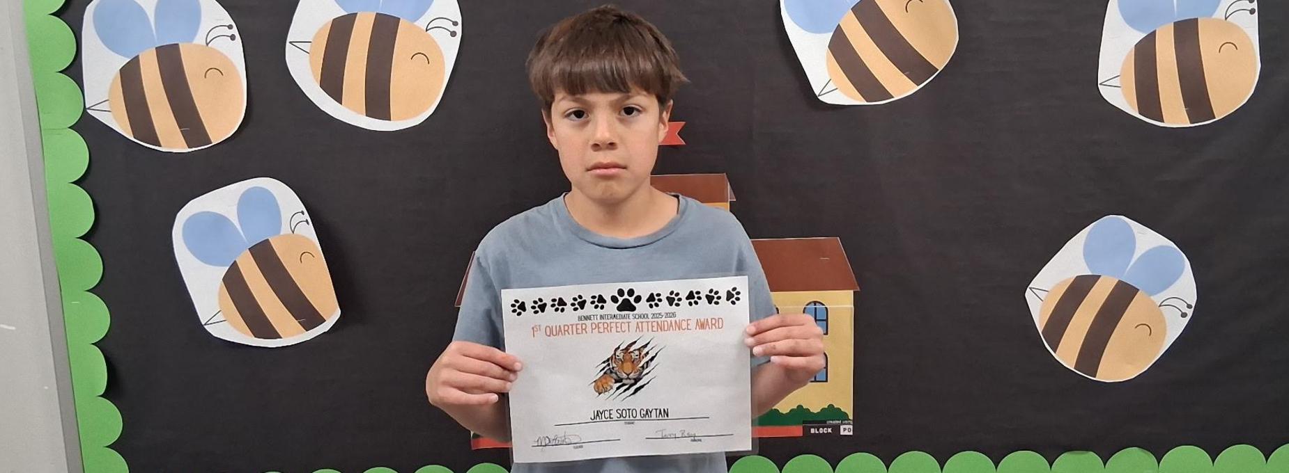 A boy holds a certificate in front of a bee-themed bulletin board.