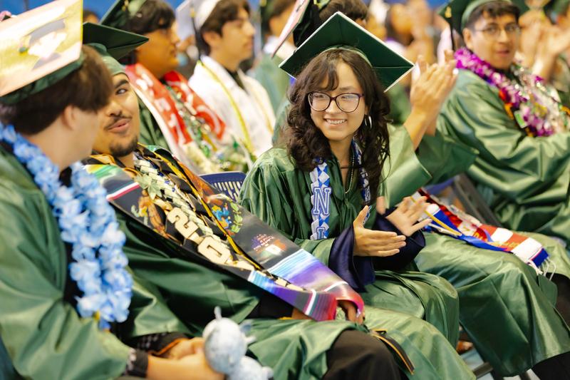 A young woman in a green cap and gown smiles and claps during a graduation ceremony while seated among fellow graduates. She wears glasses and a blue sash with the letter 