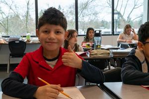 A student at Arco Iris Spanish Immersion School gives a thumbs up while working at a desk in a classroom.