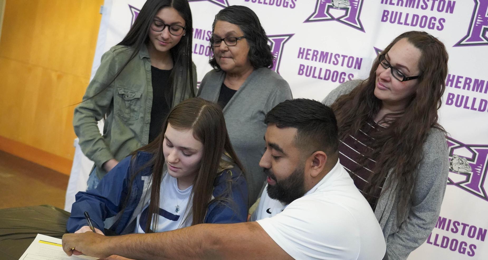 Female student signing her letter of intent with coach and family in the background.