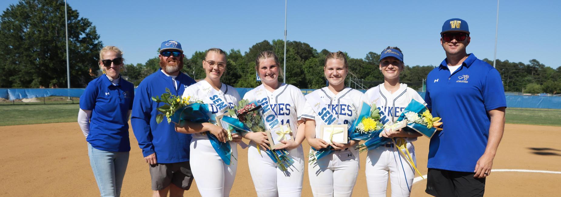 Five softball players in uniforms, holding flowers and awards on a field.