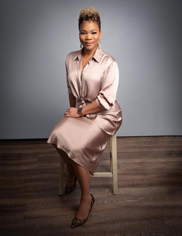 A woman in a satin dress sitting on a stool with a neutral background.