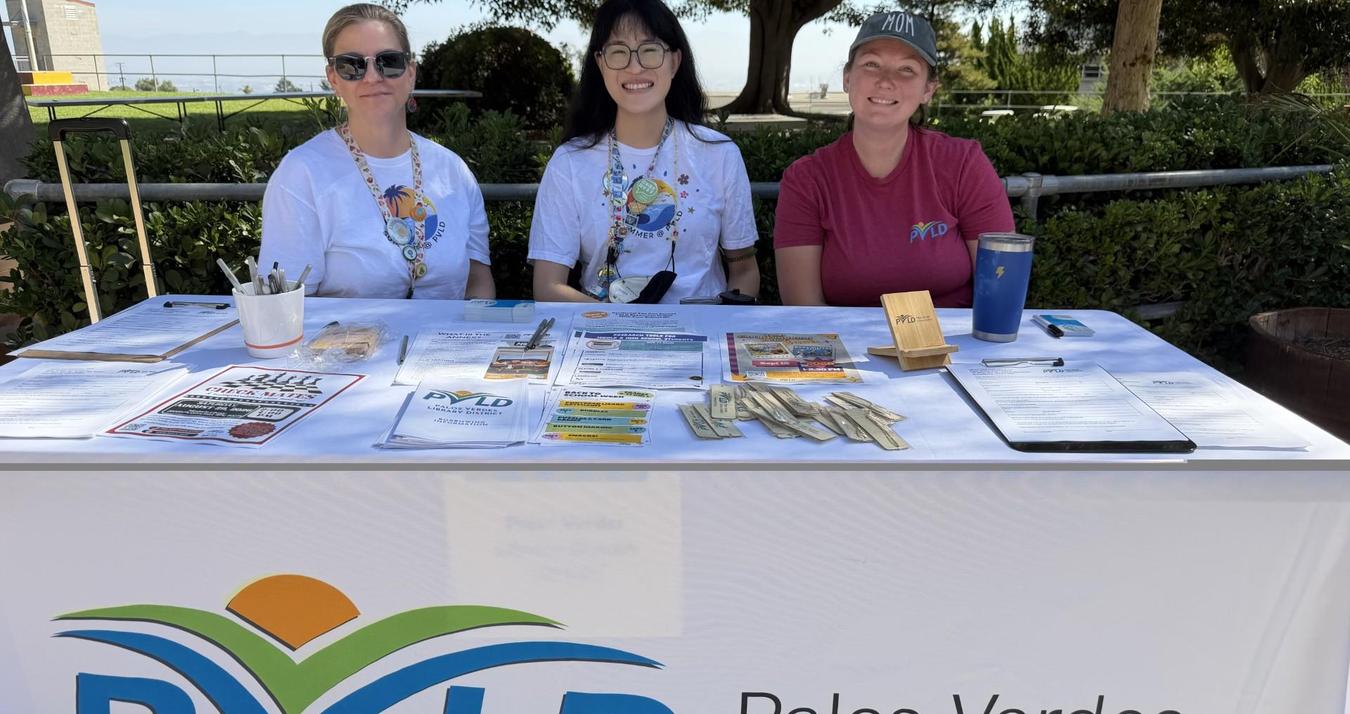 Three women smile behind a table covered with documents and promotional materials.