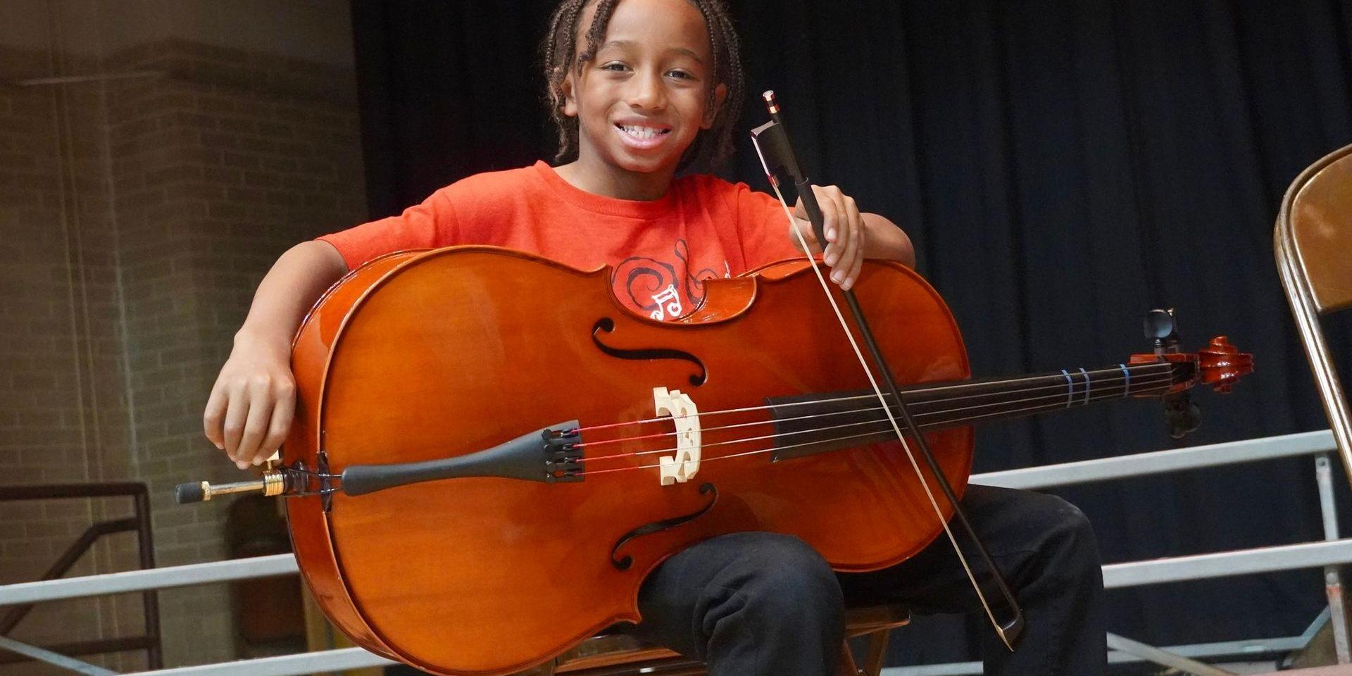 A smiling child holding a large cello while seated on a chair.