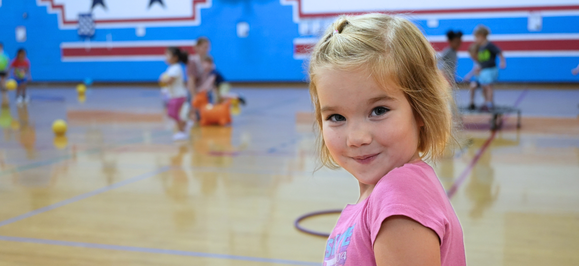Smiling girl in a gym with children playing in the background.