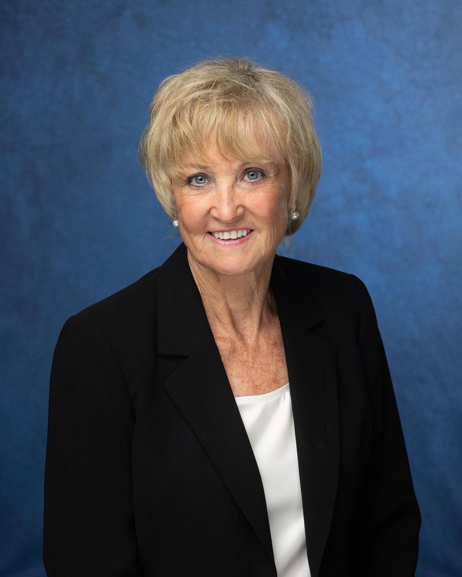 Smiling woman with short hair in a black blazer and white shirt against a blue backdrop.