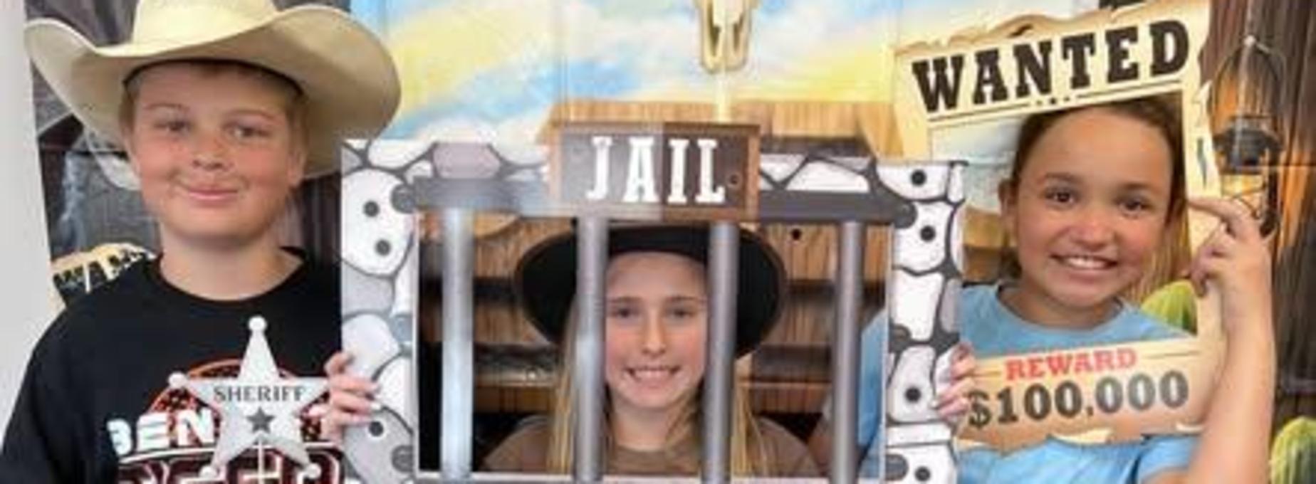 Three kids posing with props, one in a hat behind a fake jail cell frame.
