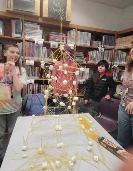 Children gather around a tall spaghetti and marshmallow structure on a table in a library.