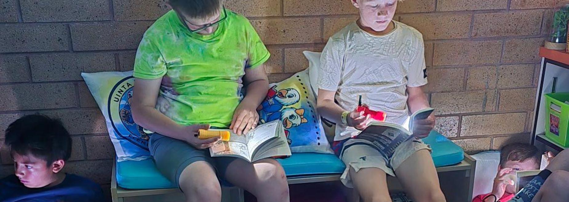 Boys sitting with books in a small classroom, colorful cushions visible around them.