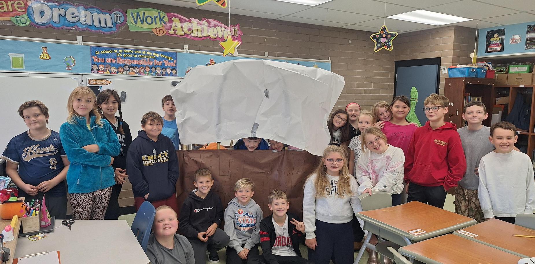 Classroom full of children gathering around a large paper structure.