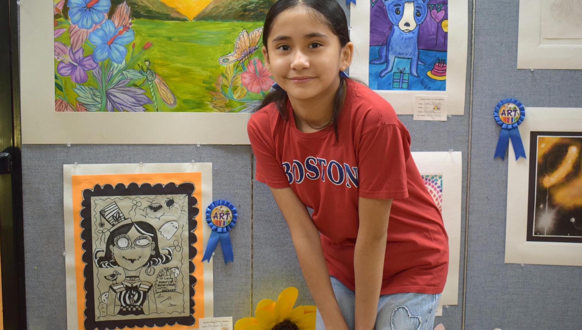 Girl in a red shirt kneeling by framed artwork with awards on a gray wall.