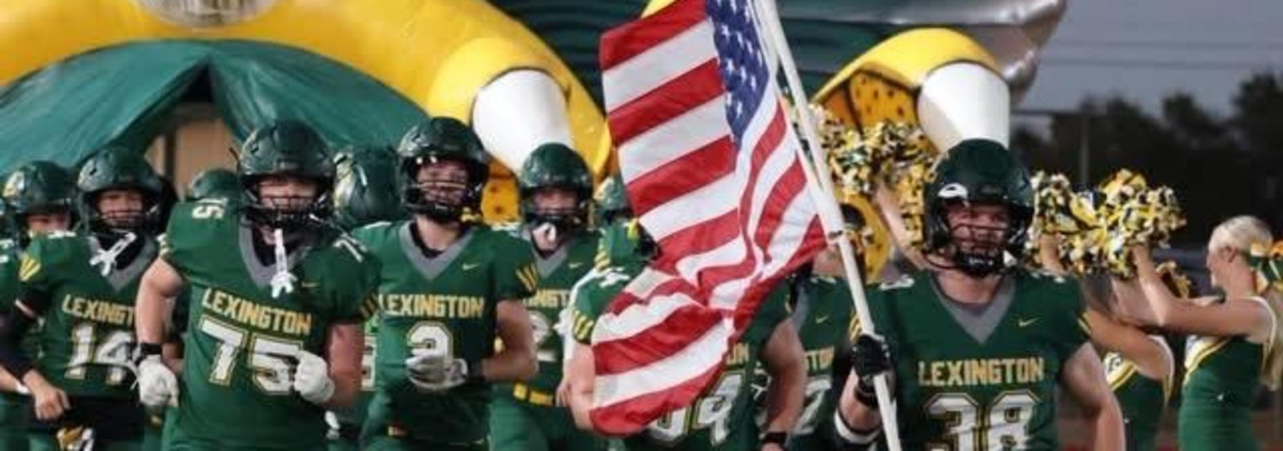 Football players running with a flag and team banners at a sports event.