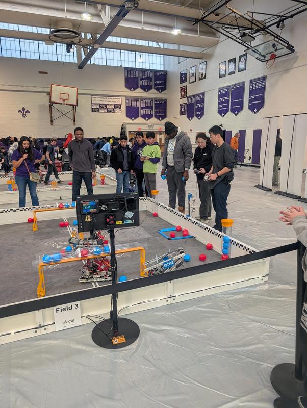 Students observing a robotics competition in a gymnasium, with robots on a field.