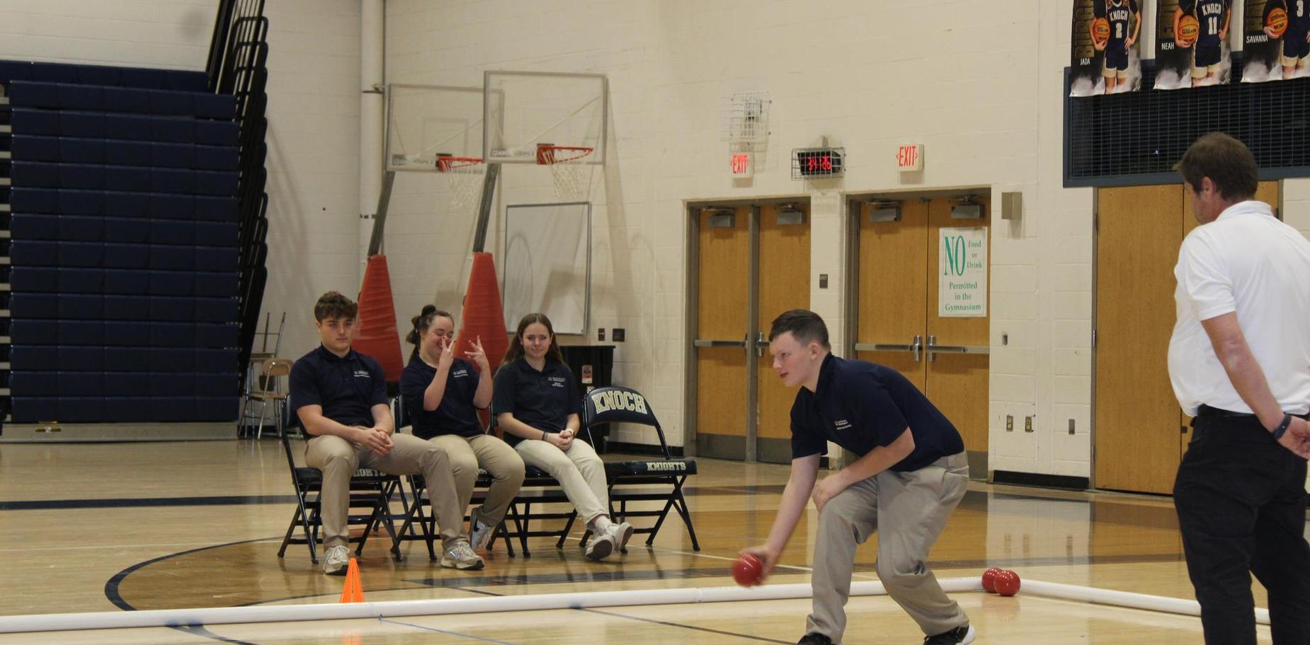 A male student rolls a red ball towards a target while others watch on bleachers.