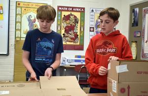 Two boys assembling cardboard boxes in a classroom.
