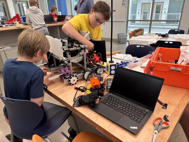 Two students collaborate at a cluttered worktable to assemble and wire a custom metal robotics project next to a laptop.