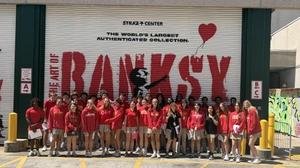 25 students in red CCC uniforms smile in front of Banksy mural