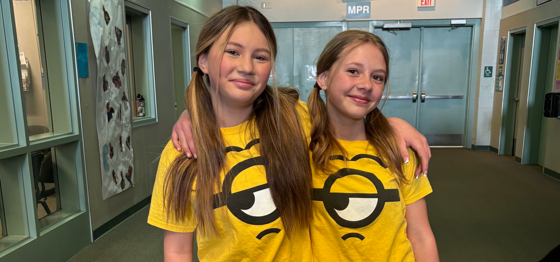 Two girls with pigtails in matching yellow shirts with a cartoon design, smiling together.