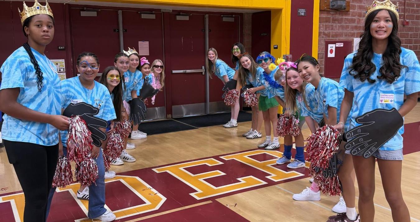 A group of girls in matching shirts pose cheerfully with pom-poms in a gymnasium.