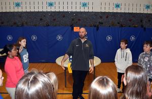 A group of children listening to a man speaking in a gymnasium.