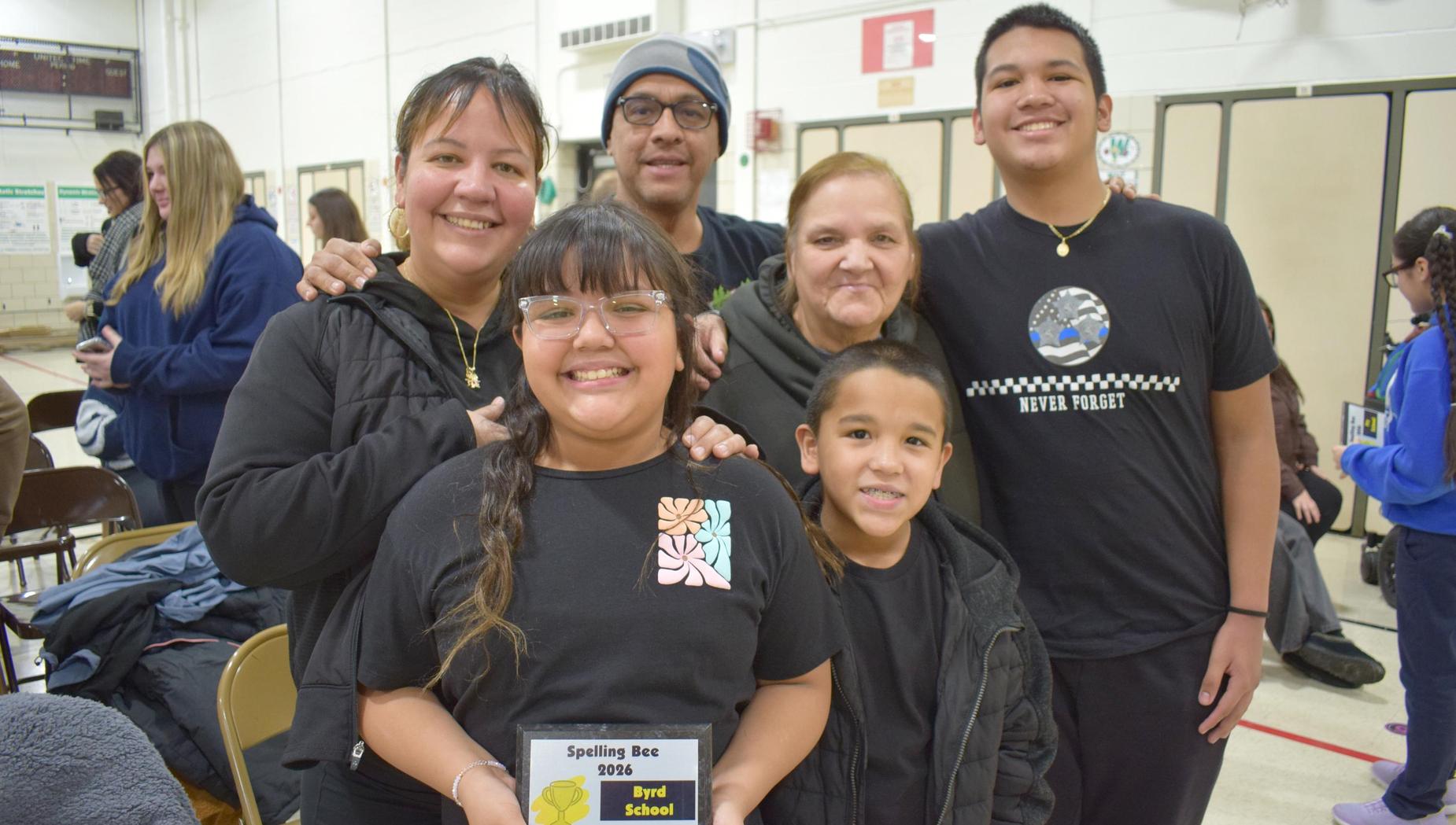 A family of six smiles together in shirts, with a girl in the front holding a spelling bee award.