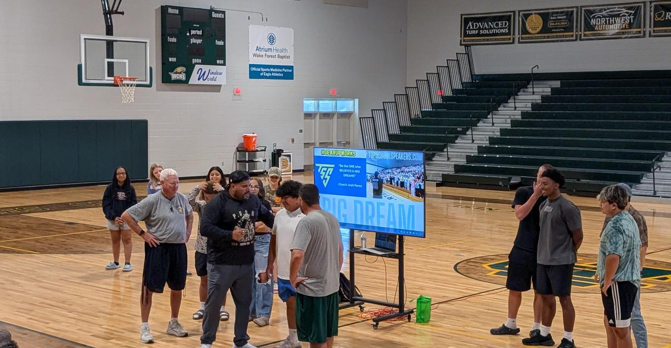 A group discusses in a gym with a screen displaying 'BIG DREAM'.