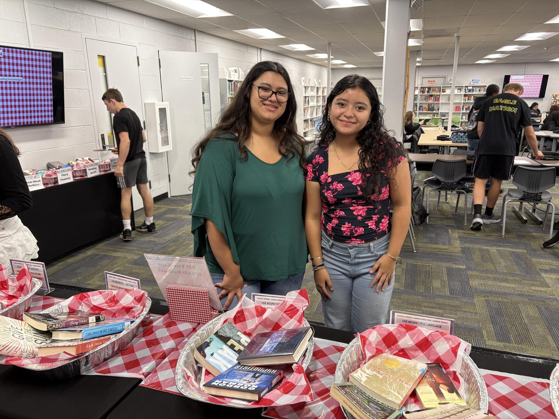 Two young women posing together with baskets of books in a library.