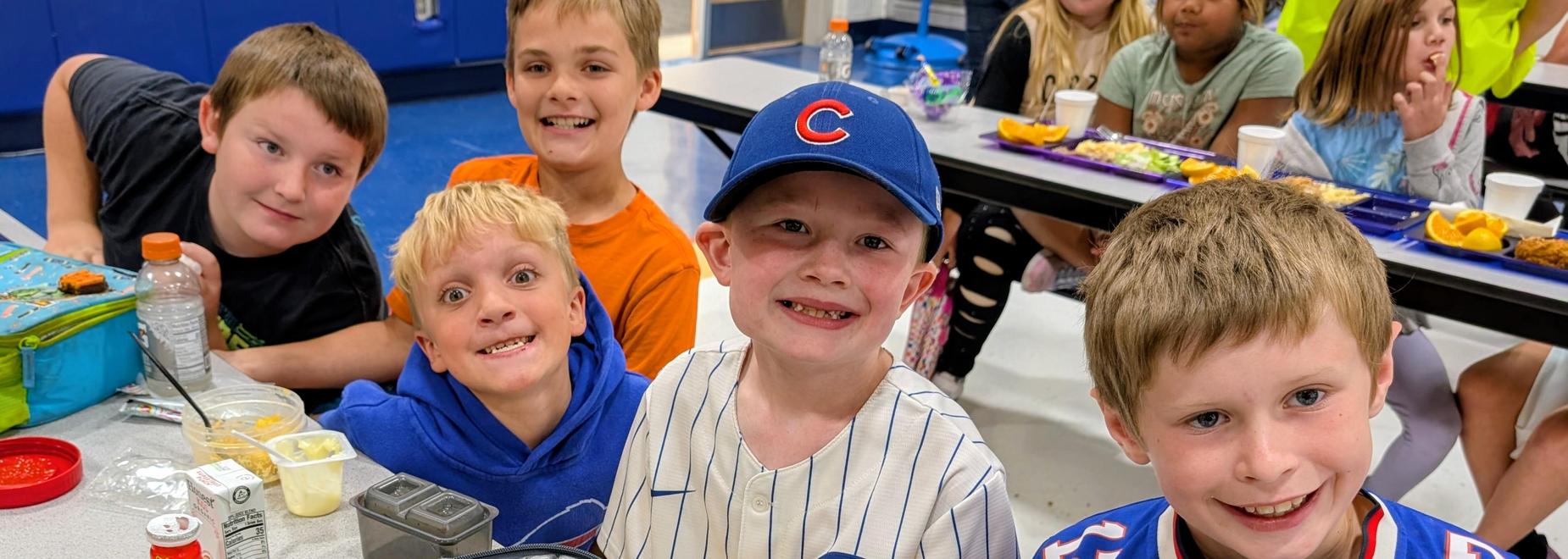 A group of five smiling children at a cafeteria table with food and drinks.