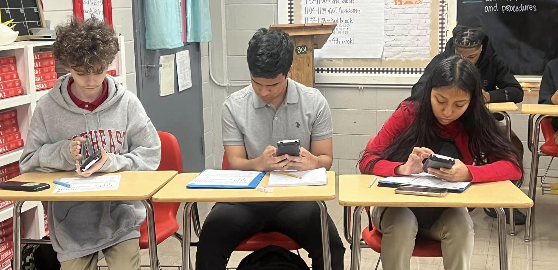 Three students focused on their phones while sitting at desks in a classroom.