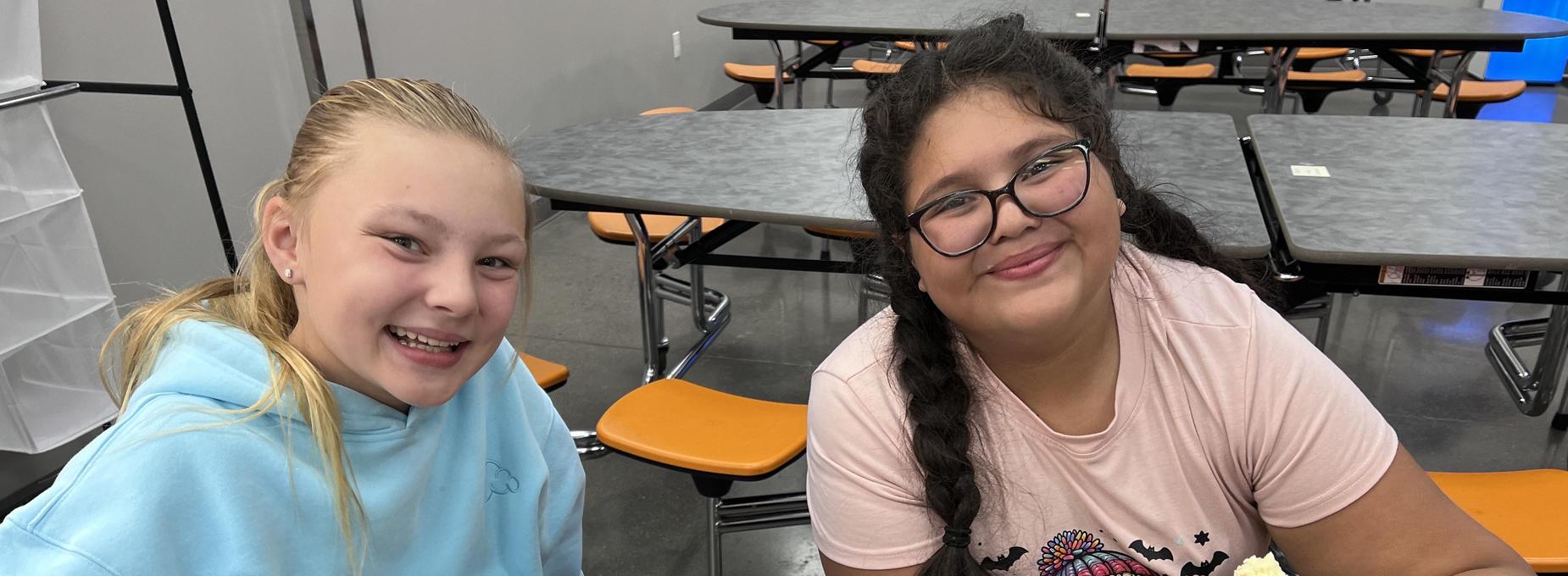 Two girls smile while enjoying donuts at a school cafeteria table.