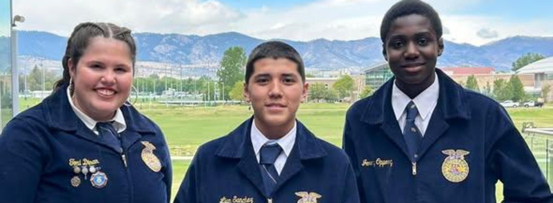 Three students in blue jackets pose together with mountains in the background.