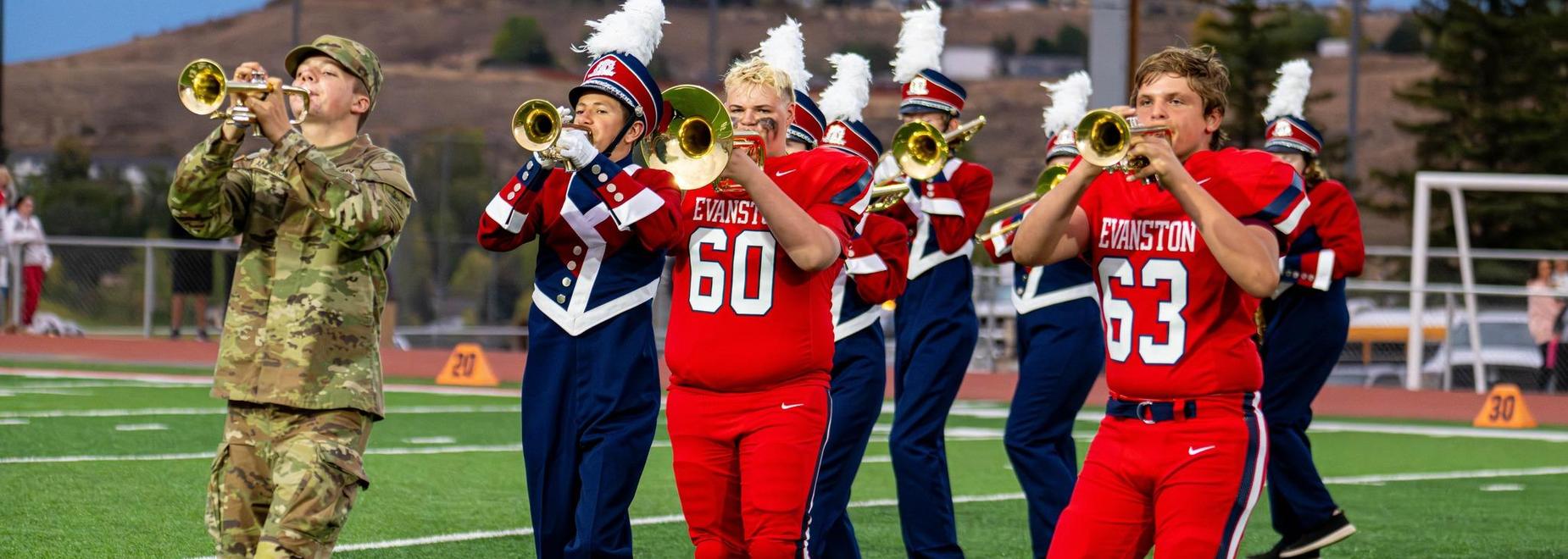 A military member and marching band members in uniforms play trumpets on a field.