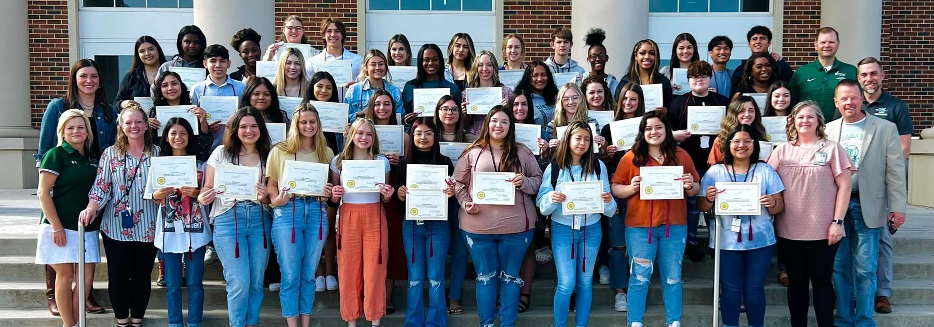 large group of teens holding certificates on stairs to campus entrance